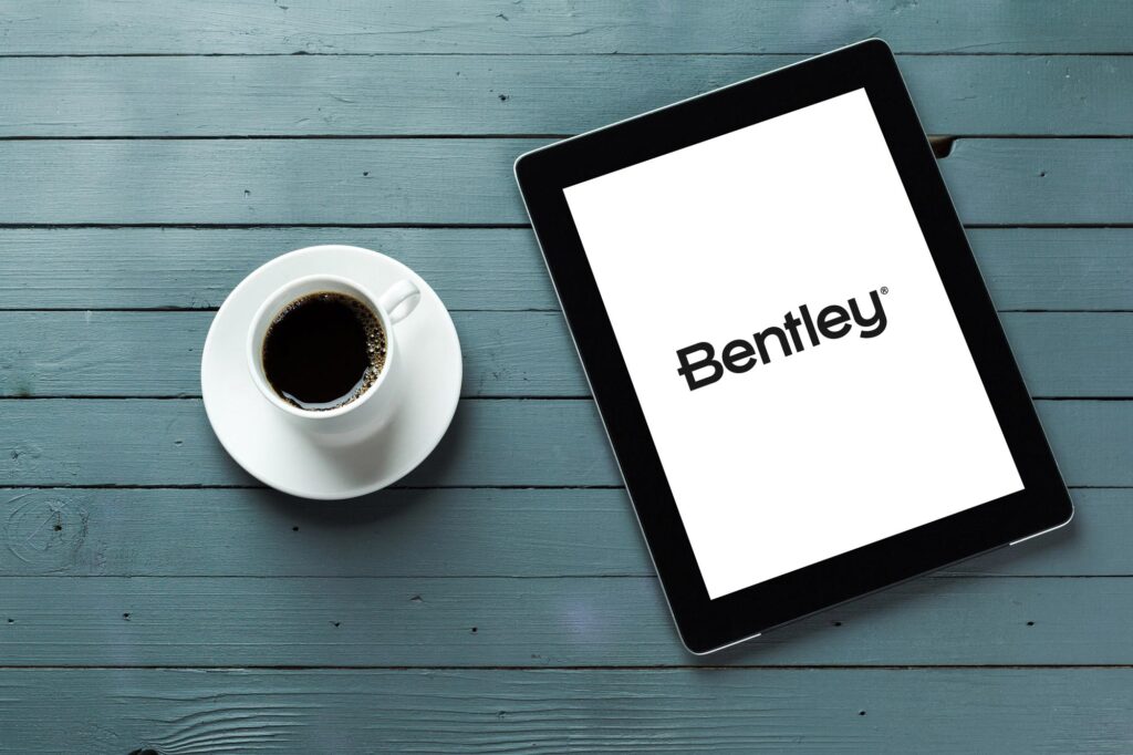 Coffee cup to the left of a tablet with the Bentley logo displaying on the tablet on blue table | Bentley Systems | Infrastructure Engineering Software Company Coffee cup to the left of a tablet with the Bentley logo displaying on the tablet on blue table