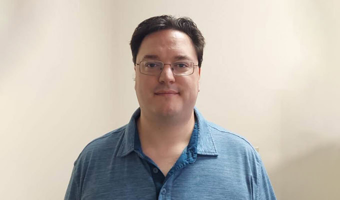 A person with short dark hair, glasses, and a blue collared shirt stands against a plain background.