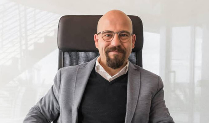 A man with glasses, a beard, and a mustache is seated in an office chair wearing a gray blazer, black sweater, and white shirt.